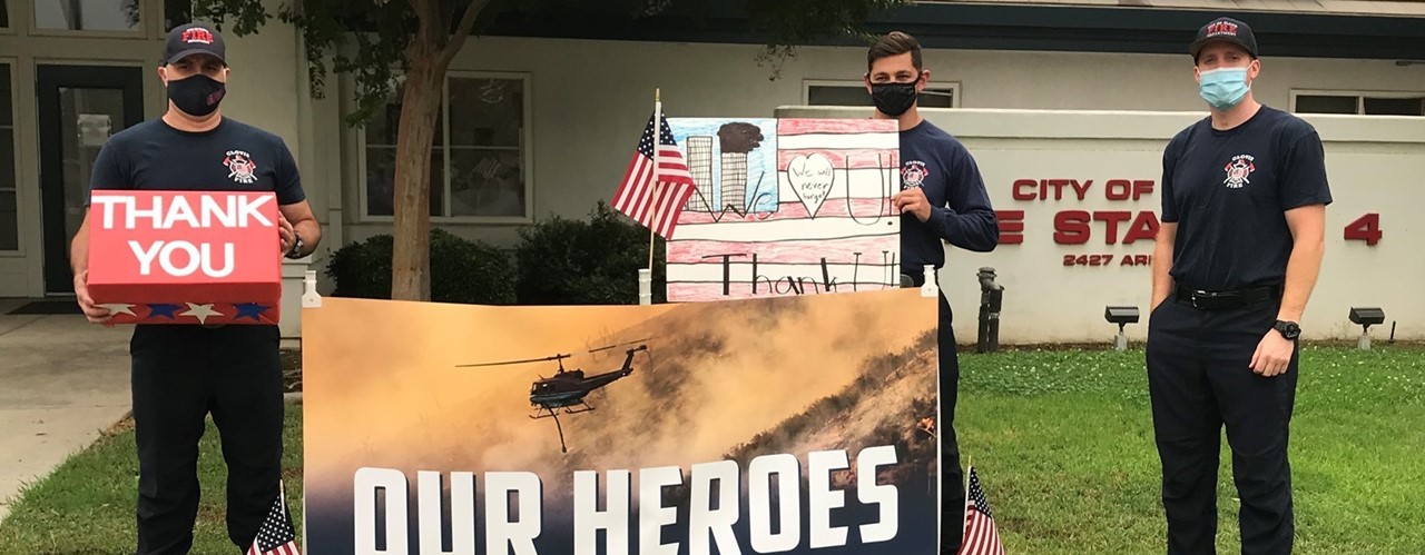 Local firefighters standing around a sign reading &#34;OUR HEROES&#34; holding pictures and a box